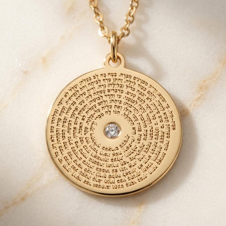 May include: A gold-toned circular pendant necklace featuring a central clear round gemstone. The surface of the pendant is engraved with dense Hebrew text arranged in concentric circles. The pendant hangs from a matching gold-toned link chain.