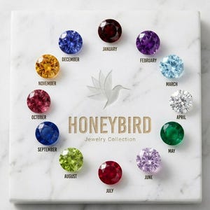 May include: A square white marble display board featuring twelve round faceted birthstones arranged in a circle. Each stone is labelled with a month from January to December. The centre displays a hummingbird logo and the text HONEYBIRD Jewelry Collection.