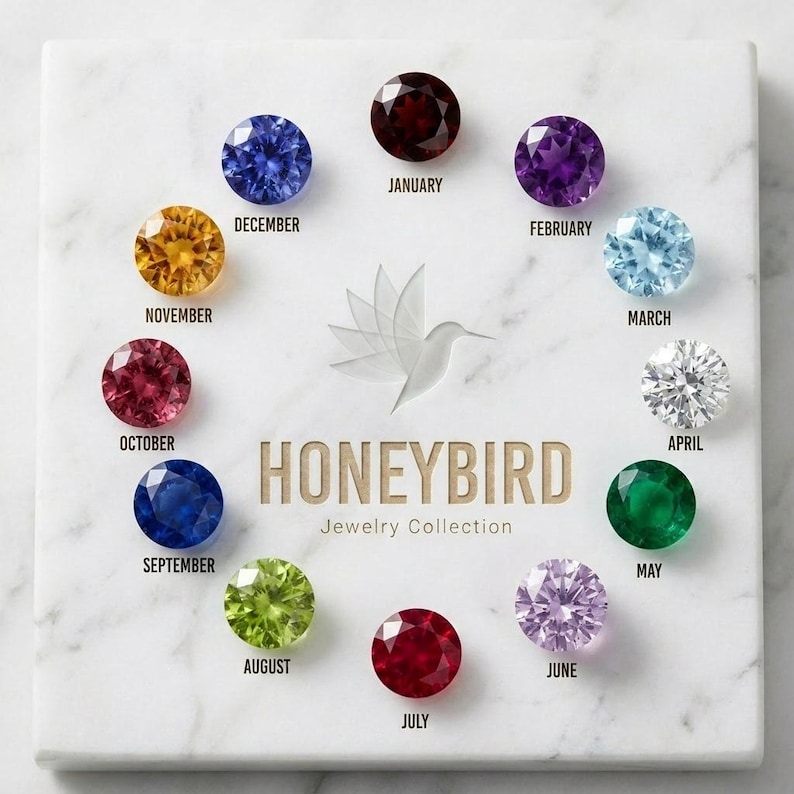 May include: A square white marble display board featuring twelve round faceted birthstones arranged in a circle. Each stone is labelled with a month from January to December. The centre displays a hummingbird logo and the text HONEYBIRD Jewelry Collection.