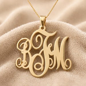 May include: A gold-colored monogram necklace with the initials BFM in a cursive font. The pendant hangs from a delicate gold chain. The necklace is displayed on a beige, textured surface.