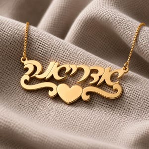 May include: A gold-colored necklace with a pendant featuring Hebrew script and a heart-shaped accent. The necklace is displayed on a textured, neutral-toned fabric, highlighting the jewelry's design and color.