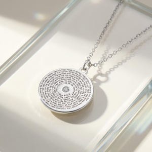 May include: A silver-toned circular pendant necklace featuring a delicate chain and a round charm engraved with dense Hebrew text. A single small, clear, round-cut gemstone is set in the centre of the pendant, surrounded by the intricate inscription.