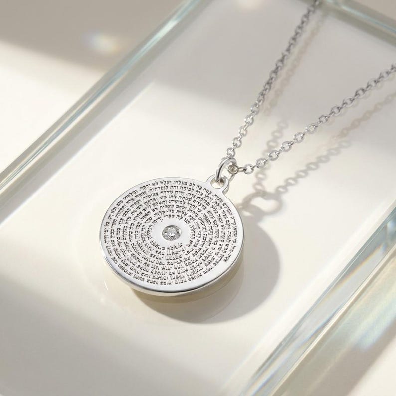 May include: A silver-toned circular pendant necklace featuring a delicate chain and a round charm engraved with dense Hebrew text. A single small, clear, round-cut gemstone is set in the centre of the pendant, surrounded by the intricate inscription.