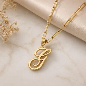 May include: A gold-coloured necklace featuring a large cursive letter "G" pendant. The chain has a paperclip link design. The necklace is displayed on cream-coloured satin fabric and a marble surface.