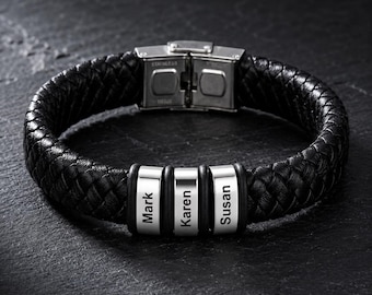 Op de afbeelding: Een zwarte gevlochten leren armband met een zilverkleurige roestvrijstalen sluiting en drie cilindervormige zilverkleurige kralen. Elke kraal is gegraveerd met een naam, namelijk Mark, Karen en Susan, gerangschikt in een rij op de armband.