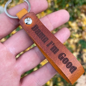 Op de afbeelding: Een bruine leren sleutelhanger met een zilveren metalen ring en een zilveren klinknagel. De leren riem is voorzien van de tekst "Damn I'm Good" in een donker, vet lettertype. De sleutelhanger heeft een getextureerd oppervlak en een vouwontwerp.