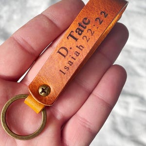 Può includere: Un portachiavi in pelle color cuoio con un anello in metallo color ottone e una chiusura a vite. Il cinturino in pelle presenta il testo inciso D. Tate e Isaiah 22:22 in un carattere scuro e leggibile, con una superficie testurizzata e ripiegata.