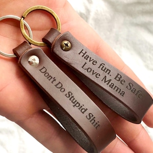 Puede incluir: Dos llaveros de cuero marrón oscuro con anillas de metal. Un llavero está grabado con el texto "Don't Do Stupid Shit" y el otro con "Have fun. Be Safe. Love Mama." Cada correa está asegurada con un pequeño tornillo de metal.