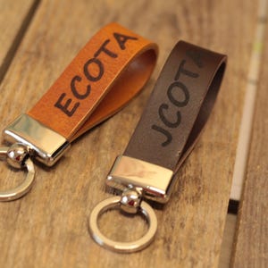 Op de afbeelding: Twee lederen sleutelhangers met zilverkleurige metalen hardware en ronde ringen. De ene sleutelhanger is van bruin leer met de tekst ECOTA in het zwart gedrukt, de andere is van donkerbruin leer met de tekst JCOTA in het zwart op de band.