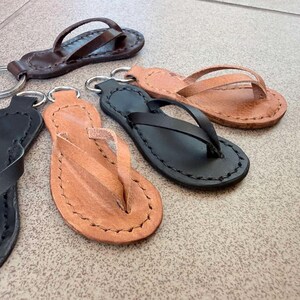 Könnte beinhalten: Vier Schlüsselanhänger in Form von Flip-Flops aus Leder in Schwarz und Hellbraun. Jeder Anhänger hat die Form einer Miniatur-Sandale mit sichtbaren Nähten an den Rändern und einem Metallring an der Ferse zur Befestigung von Schlüsseln.