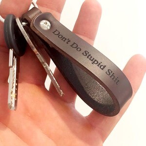 Puede incluir: Un llavero de cuero marrón oscuro con un cierre de tornillo de metal plateado y el texto grabado Don't Do Stupid Shit. La correa de cuero está doblada en un bucle y unida a una anilla de metal plateado que sostiene dos llaves de metal.