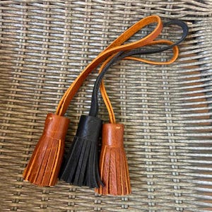 May include: Three leather tassels attached to long, thin leather straps. Two of the tassels are a warm, medium-brown tan color, and one central tassel is a solid black. Each tassel features multiple vertical fringe strips and a smooth top.