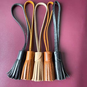 Pode incluir: Cinco borlas de couro dispostas em fila, com um gradiente de cor que vai do preto ao castanho claro, bege, castanho médio e preto novamente. Cada borla tem uma alça de couro longa e fina no topo e uma base cilíndrica com franjas verticais.