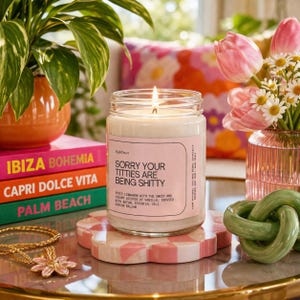 May include: A burning candle in a glass jar with a pink label that reads "Sorry Your Titties Are Being Shitty." The candle is on a pink and white checkered coaster. Other items include a stack of colourful books, flowers, and a gold necklace.
