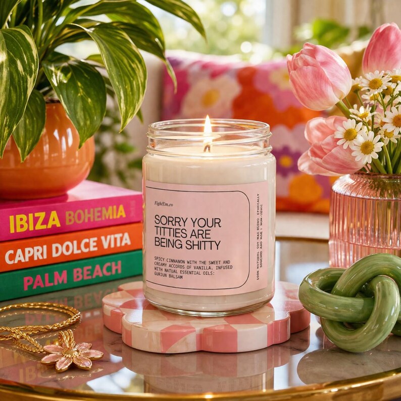 May include: A burning candle in a glass jar with a pink label that reads "Sorry Your Titties Are Being Shitty." The candle is on a pink and white checkered coaster. Other items include a stack of colourful books, flowers, and a gold necklace.