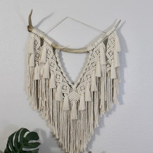 May include: A macrame wall hanging with a diamond pattern and tassels, hanging from a pair of antlers.