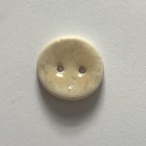 Op de afbeelding: Een ronde, gebroken witte knoop met twee gaatjes. De knoop heeft een licht onregelmatige vorm en een glanzende afwerking. De kleur is een warme, crèmekleurige tint met subtiele variaties.