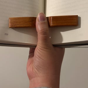 May include: A wooden bookmark is inserted into a book. The bookmark is a rectangular shape with a warm, reddish-brown color. The number 222 is visible on the page. The book is held open.