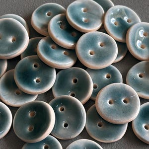 May include: A collection of round ceramic buttons featuring a glossy teal and blue glaze with a smooth finish. Each button has two central holes for attachment and a light-colored, unglazed edge, creating a contrast with the dark blue center.