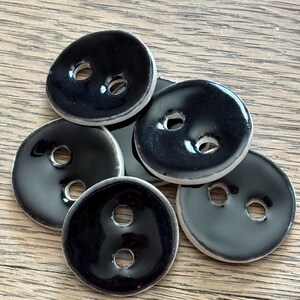 May include: A collection of five round, two-hole buttons featuring a glossy black finish on the top surface and a contrasting white edge. Each button has a slightly irregular, organic shape with a smooth, reflective surface texture.