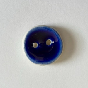 Puede incluir: Un botón redondo de cerámica con un centro azul oscuro brillante y un borde blanco. El botón tiene dos pequeños agujeros para coser. El botón está sobre un fondo blanco.