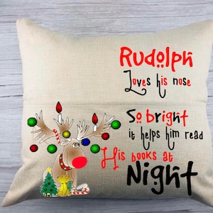 May include: A beige pillow with a red and black illustration of Rudolph the Red-Nosed Reindeer. The text on the pillow reads "Rudolph loves his nose so bright it helps him read his books at night".