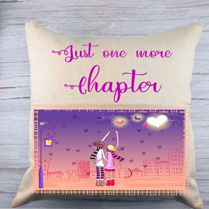 Op de afbeelding: Een decoratief kussen met een beige stoffen hoes en een paarse en roze grafische afbeelding van een stel dat in een stad staat met harten in de lucht. De tekst "Just one more chapter" is in paars op het kussen gedrukt.