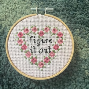 May include: A cross-stitch embroidery hoop with the words "figure it out" in black script. The design features a heart-shaped floral border in pink and green. The hoop is made of wood and is approximately 6 inches in diameter.
