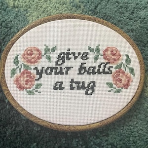 May include: Oval cross-stitch embroidery in a wooden frame. The text "give your balls a tug" is stitched in black, surrounded by pink roses and green leaves. The background is white.