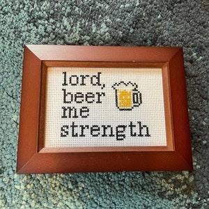 Op de afbeelding: Een ingelijst kruissteek met de tekst "Heer, geef me bier" en een gele bierpul-afbeelding.