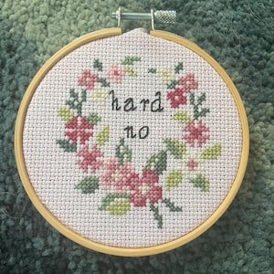 May include: A cross-stitch embroidery hoop with the words "hard no" in black lettering. The text is surrounded by a floral wreath in shades of pink, green, and white. The hoop is made of light-colored wood.