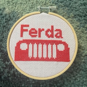 May include: A cross-stitch embroidery hoop featuring the word "Ferda" in red block letters above a red Jeep-style grille design. The embroidery is on white fabric, set in a wooden hoop.