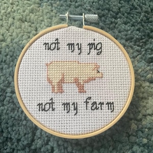 May include: A cross-stitch embroidery hoop featuring a pig design in tan and brown. The text "not my pig" and "not my farm" are stitched in black above and below the pig. The hoop is made of wood.