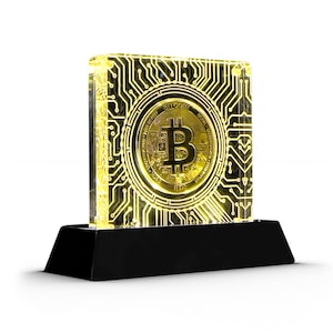 Puede incluir: Una moneda de criptomoneda Bitcoin de color dorado se muestra en un bloque de acrílico transparente con un diseño de placa de circuito. El bloque está iluminado desde adentro, proyectando un brillo amarillo cálido. El bloque se asienta sobre una base negra.
