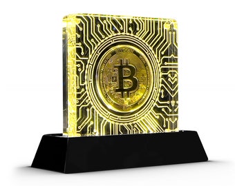 May include: A gold-coloured Bitcoin cryptocurrency coin is displayed in a clear acrylic block with a circuit board design. The block is illuminated from within, casting a warm yellow glow. The block sits on a black base.