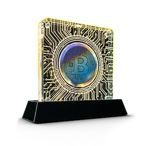 May include: A gold-colored Bitcoin coin encased in a clear acrylic block with a circuit board design. The block sits on a black base.