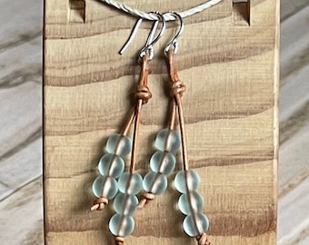 May include: A pair of dangle earrings with light blue glass beads and brown leather cord. The earrings are displayed on a wooden stand.