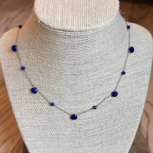 May include: A minimalist necklace with a fine chain, adorned with evenly spaced, deep blue beads. The beads are oval-shaped and create a simple yet elegant design. The necklace is displayed on a neutral-toned bust, showcasing its delicate nature.
