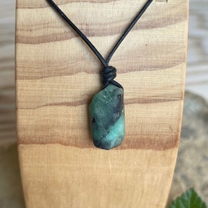 May include: A dark green emerald gemstone pendant on a black leather cord necklace. The pendant is a rough, natural shape and is hanging from a wooden display stand.