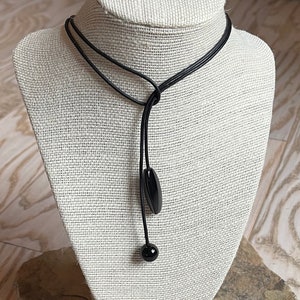 May include: A black leather choker necklace with a black bead and a black pendant. The necklace is displayed on a fabric-covered mannequin.
