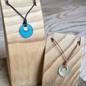 May include: Two necklaces with sea glass pendants on wooden display stands. The necklace on the left has a blue sea glass pendant and a black cord. The necklace on the right has a white sea glass pendant and a brown cord.