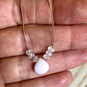 May include: Delicate necklace featuring a gold chain with small, clear beads and a pear-shaped, white pendant. The necklace is held up against a neutral background, showcasing its simple elegance. A beautiful piece of jewelry.