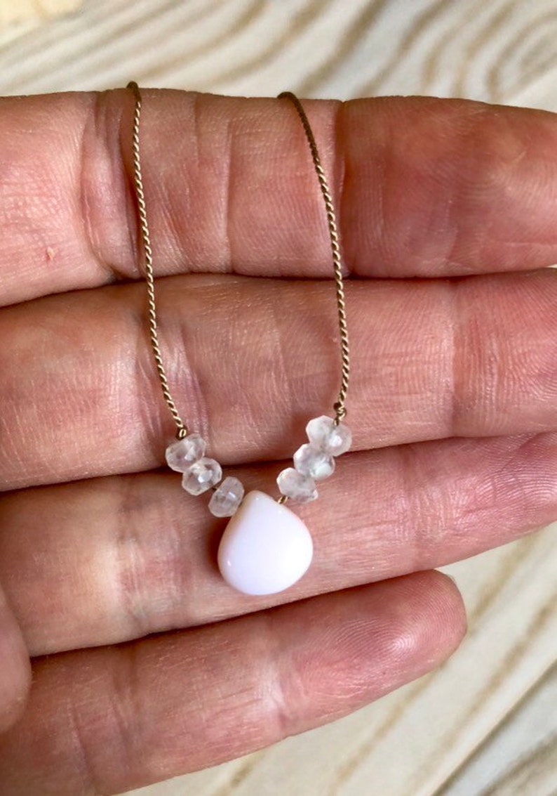 May include: Delicate necklace featuring a gold chain with small, clear beads and a pear-shaped, white pendant. The necklace is held up against a neutral background, showcasing its simple elegance. A beautiful piece of jewelry.