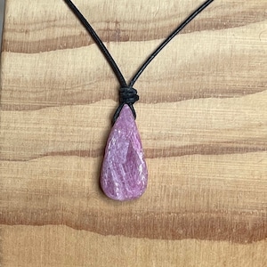 May include: A pink gemstone pendant on a black leather cord necklace. The pendant is a teardrop shape with a rough, natural finish.
