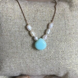 May include: A delicate necklace featuring a gold chain, freshwater pearls, and a teardrop-shaped, light blue gemstone pendant. The necklace is displayed on a textured, beige surface.