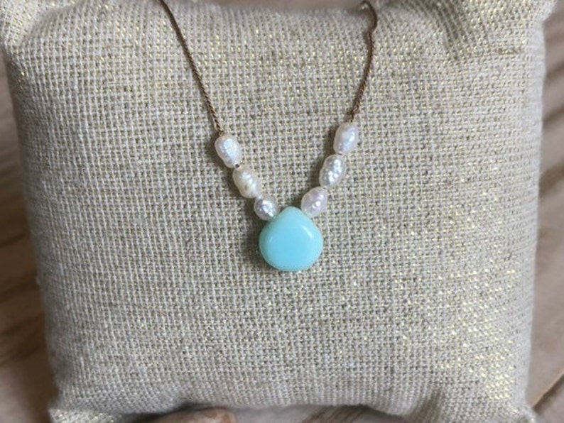 May include: A delicate necklace featuring a gold chain, freshwater pearls, and a teardrop-shaped, light blue gemstone pendant. The necklace is displayed on a textured, beige surface.