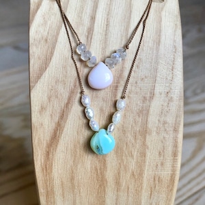 May include: Two-strand necklace with a light pink teardrop pendant and a light blue, round pendant. The top strand features small, clear beads, while the bottom strand has white pearl-like beads. The necklace is displayed on a wooden stand.