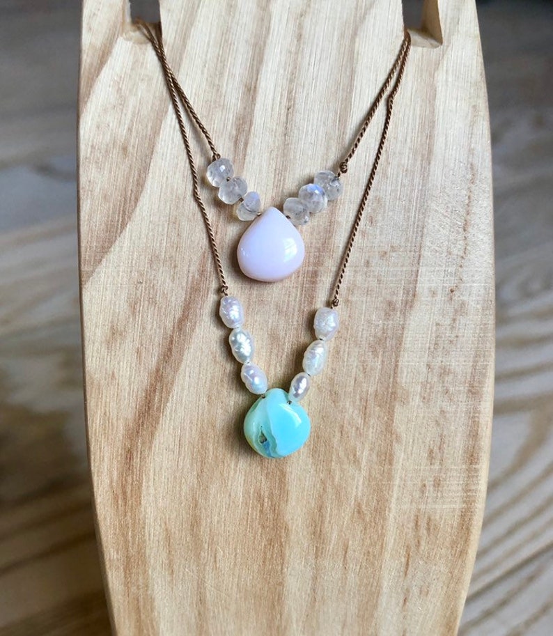 May include: Two-strand necklace with a light pink teardrop pendant and a light blue, round pendant. The top strand features small, clear beads, while the bottom strand has white pearl-like beads. The necklace is displayed on a wooden stand.