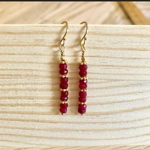 May include: A pair of gold-colored earrings with a vertical design. Each earring features a series of small, faceted, red beads separated by gold-colored spacers. The earrings hang from gold-colored ear wires.