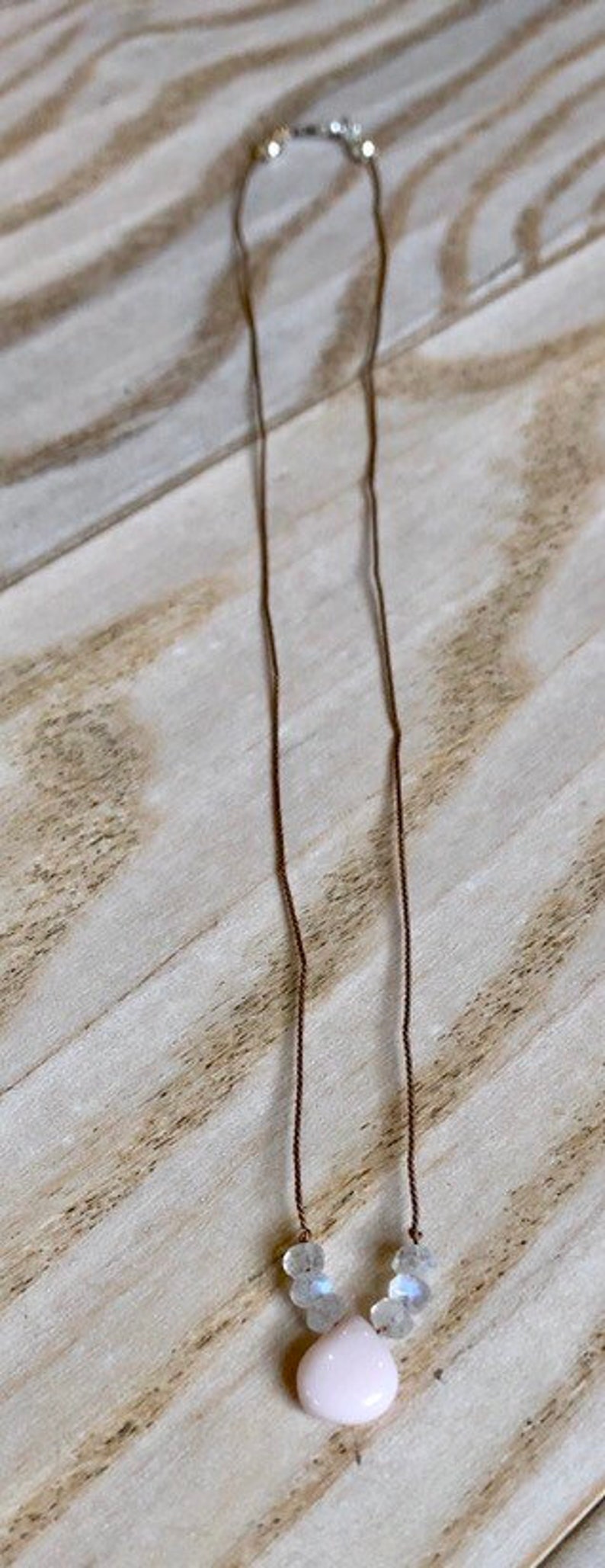 May include: A delicate necklace featuring a light pink teardrop-shaped pendant and clear, faceted beads. The necklace is strung on a thin, brown cord and has a silver clasp.
